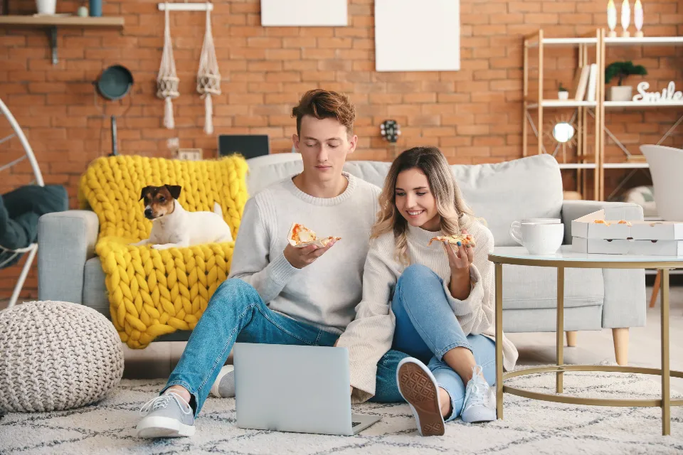 Young couple sit on a cozy living room rug with a laptop and pizza slices, a small dog resting on a yellow knit blanket behind them