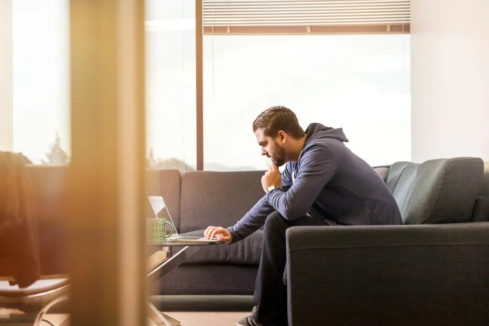Man in a navy hoodie sits on a gray sofa working on a laptop in a bright, modern furnished room with large windows.