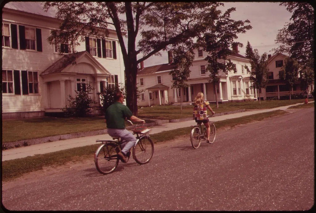 Two people cycle along a quiet tree-lined residential street past white clapboard houses with green lawns.